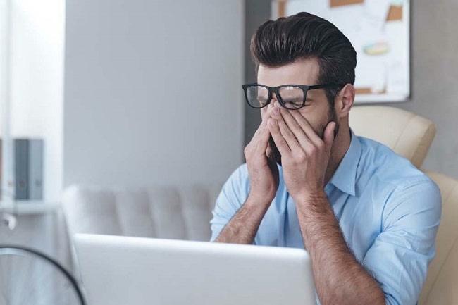 A man sits at his desk, his hands covering his face in a gesture of frustration or deep thought.