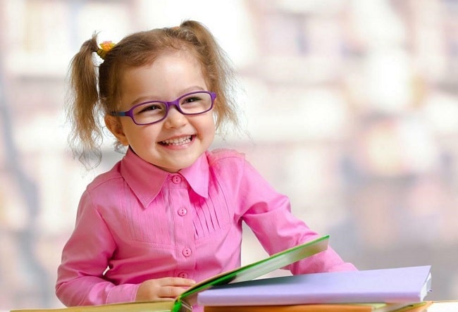 A studious little girl with glasses sits at a table surrounded by books, engrossed in her studies.