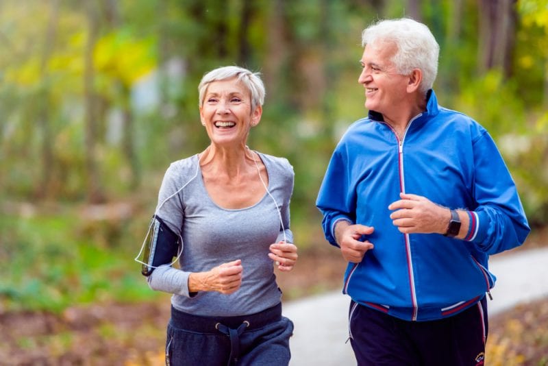 Senior man and woman smiling and running side by side in the park.